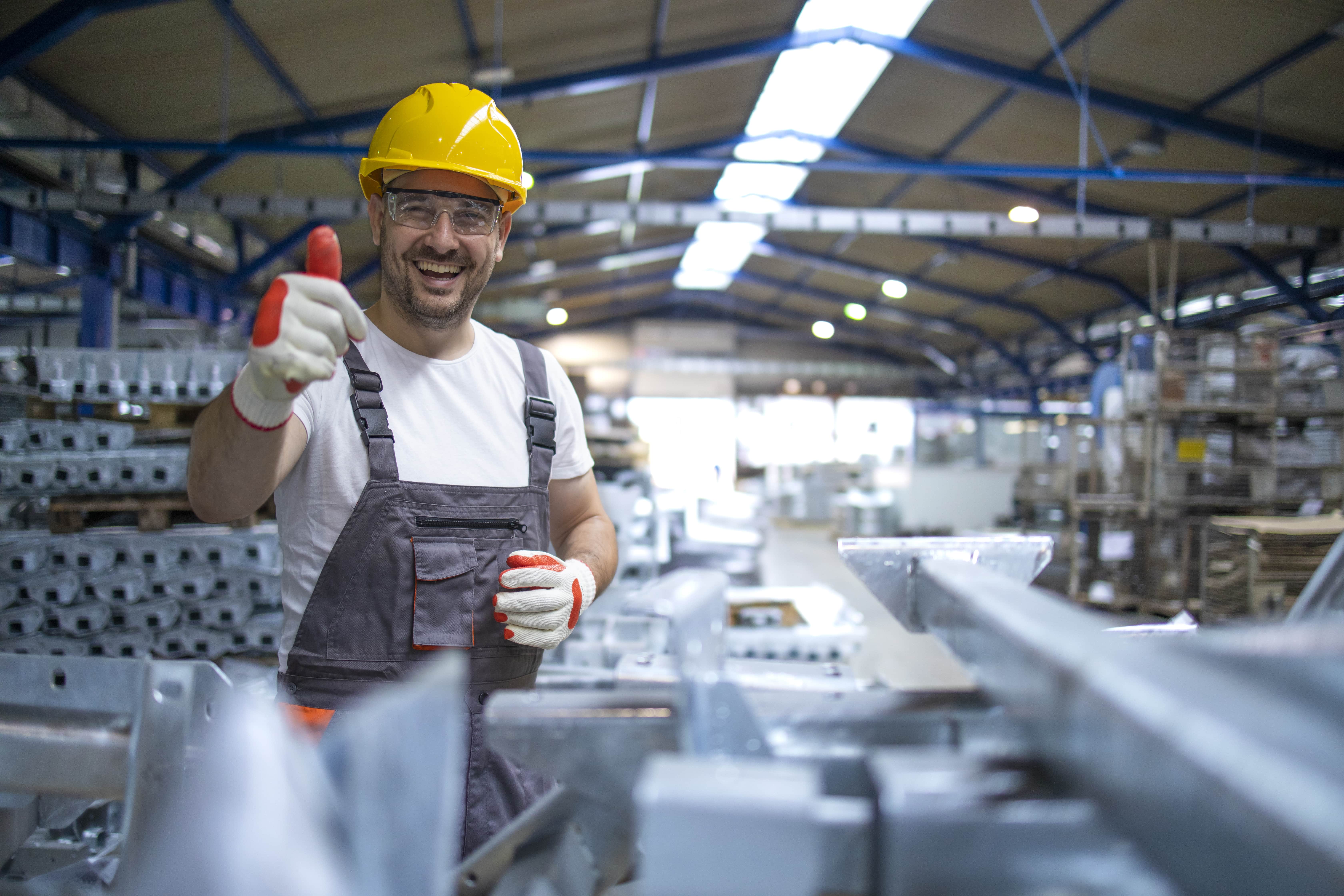 Homem branco com capacete amarelo e luvas de proteção, ambiente de fabrica fazendo o sinal de positivo.